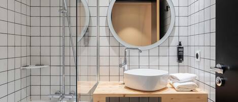 Un baño moderno con ducha de vidrio, un espejo redondo, un lavamanos blanco y un mueble de tocador de madera.