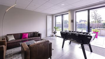 A modern living room with a sofa, armchairs, a foosball table, and large windows offering a view of the outdoors.