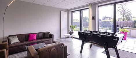 Een moderne woonkamer met een bank, fauteuils, een tafelvoetbalspel en grote ramen met uitzicht op de omgeving.