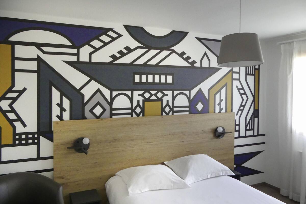 A bedroom with a wooden headboard, a bed with white linens, a black chair, and a wall with geometric patterns.