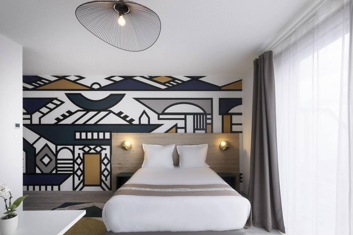 A modern hotel room with a bed, bedside lamps, a geometric wall design, and a window with curtains.