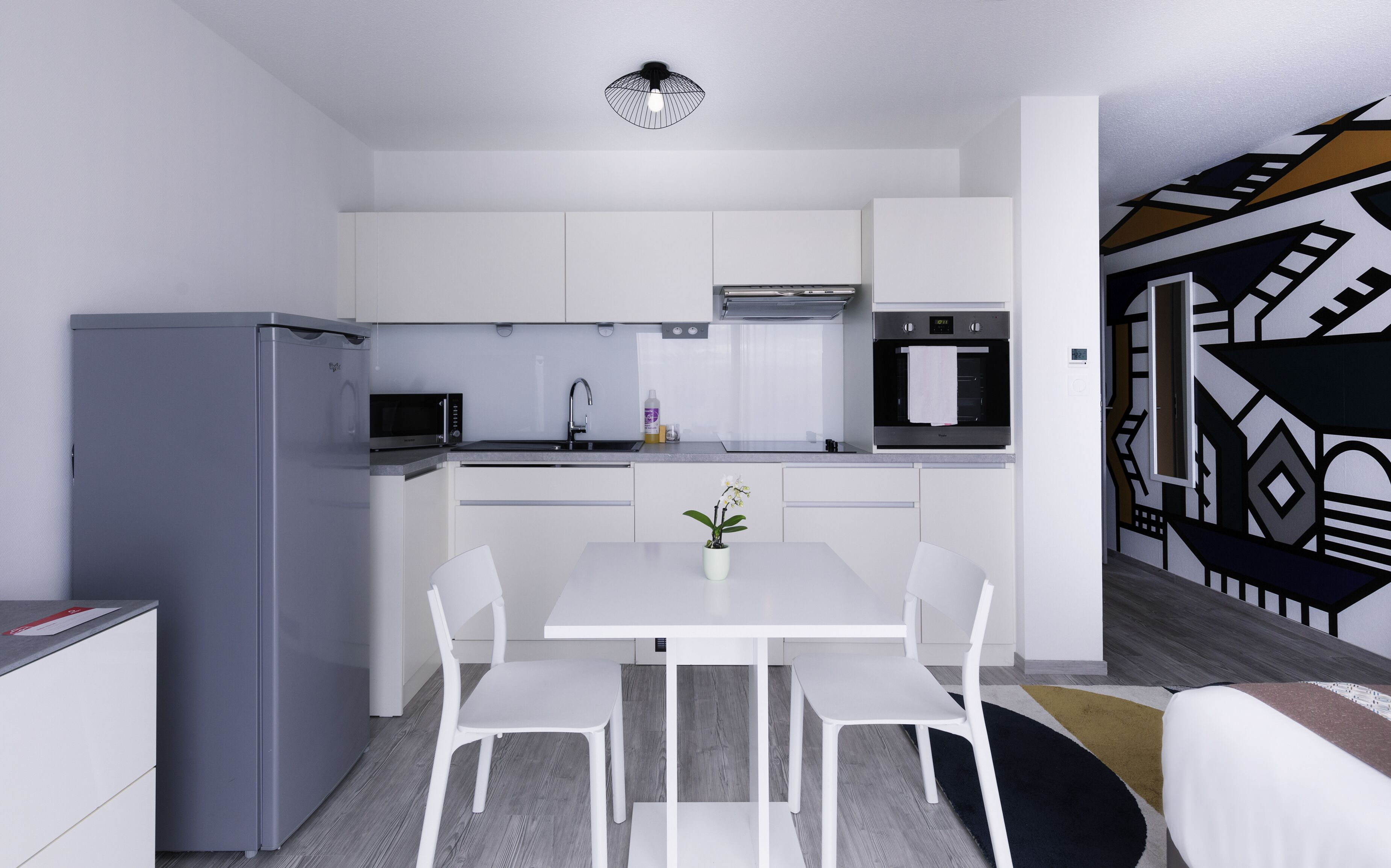 A modern kitchen with white cabinets, a grey refrigerator, a microwave, and an oven. There is a dining table with white chairs and a small potted plant.