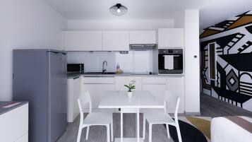 A modern kitchen with white cabinets, a grey refrigerator, a microwave, and an oven. There is a dining table with white chairs and a small potted plant.