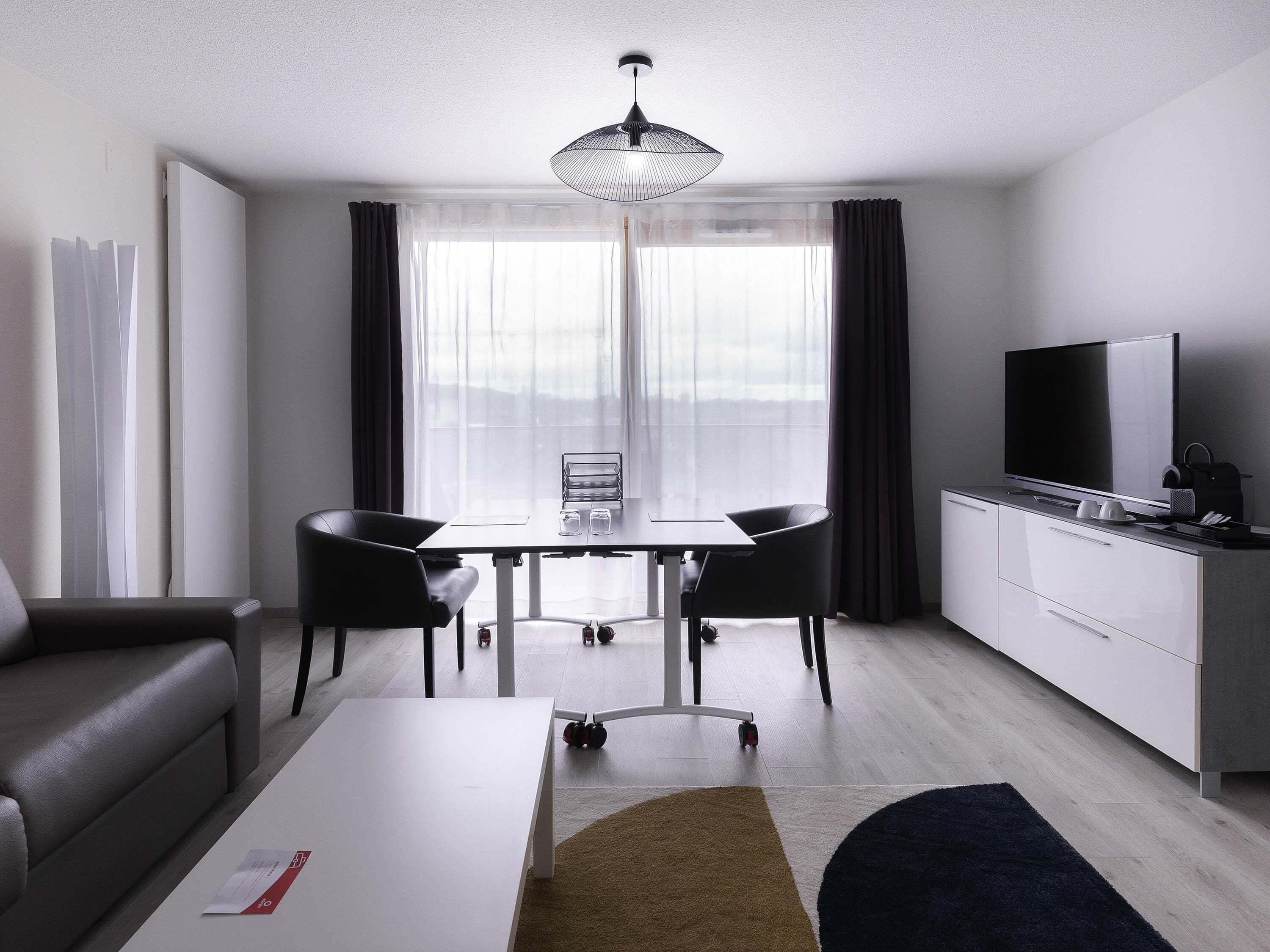 Una habitación de hotel moderna con una mesa de comedor, sillas, un sofá, un televisor y una kitchenette.