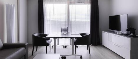 A modern hotel room with a dining table, chairs, a sofa, a TV, and a kitchenette.