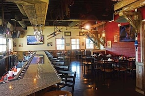 Zona de bar con una barra larga, varios tipos de asientos y una escalera que sube a un nivel superior.