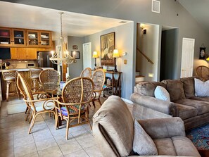 A living room with a brown sofa, wicker chairs, a dining area with a chandelier, and a kitchen in the background. - River Haven: lakefront home with dock and kayaks (McQueeney)