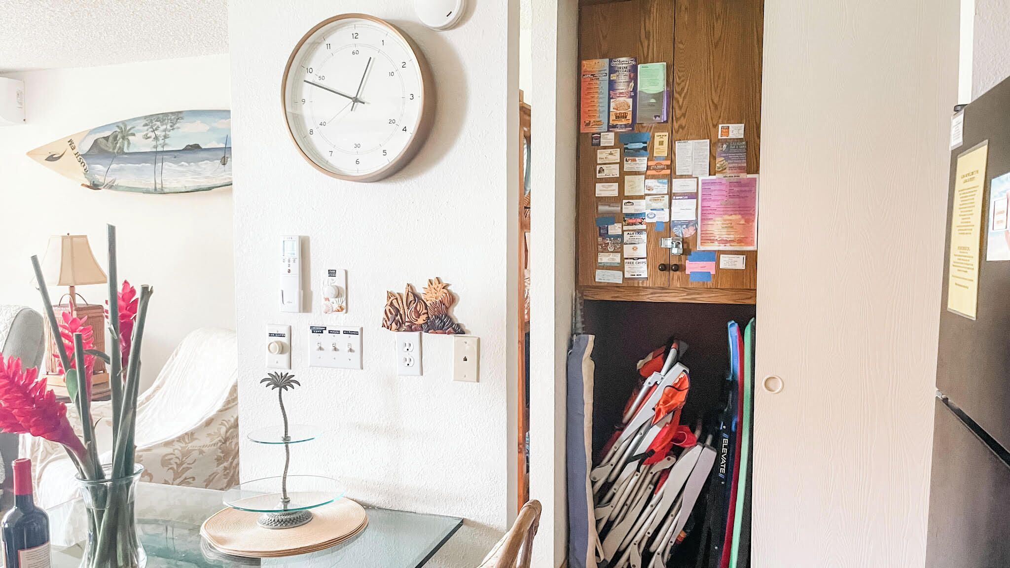 A room with a wall clock, a bulletin board with various notices, a surfboard decoration, and a table with a vase of flowers.