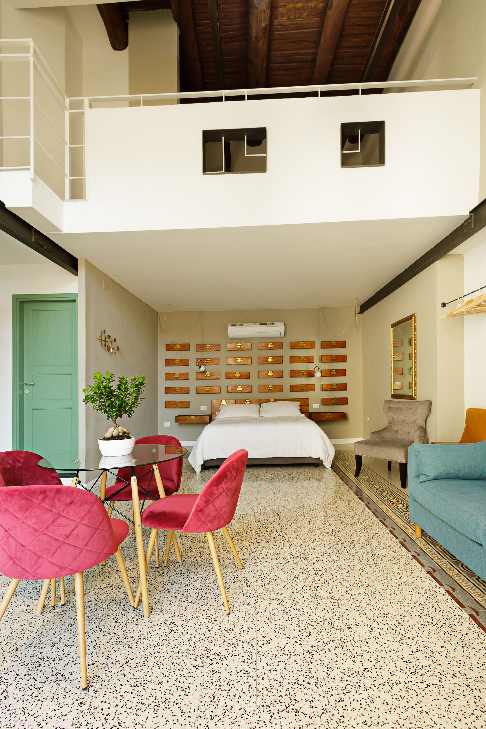 A modern hotel room with a bed, a dining area with red chairs, a small table, a potted plant, and a wall with wooden storage boxes.