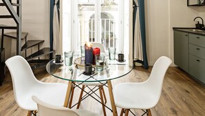 Uma área de refeições com mesa de vidro, cadeiras brancas e vista para uma varanda através de cortinas azuis.