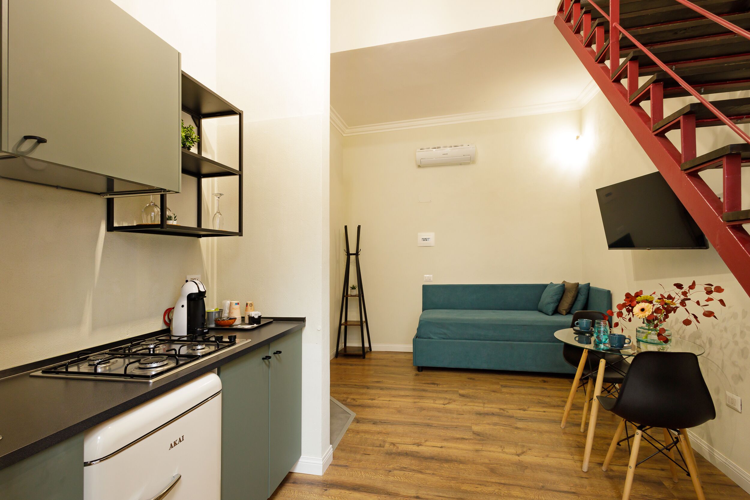 Une cuisine compacte équipée d’une cuisinière à gaz, d’un petit réfrigérateur et d’une étagère intégrée. On y trouve un coin repas avec une table et des chaises, ainsi qu’un canapé bleu à l’arrière-plan. Un escalier rouge est visible sur le côté droit.