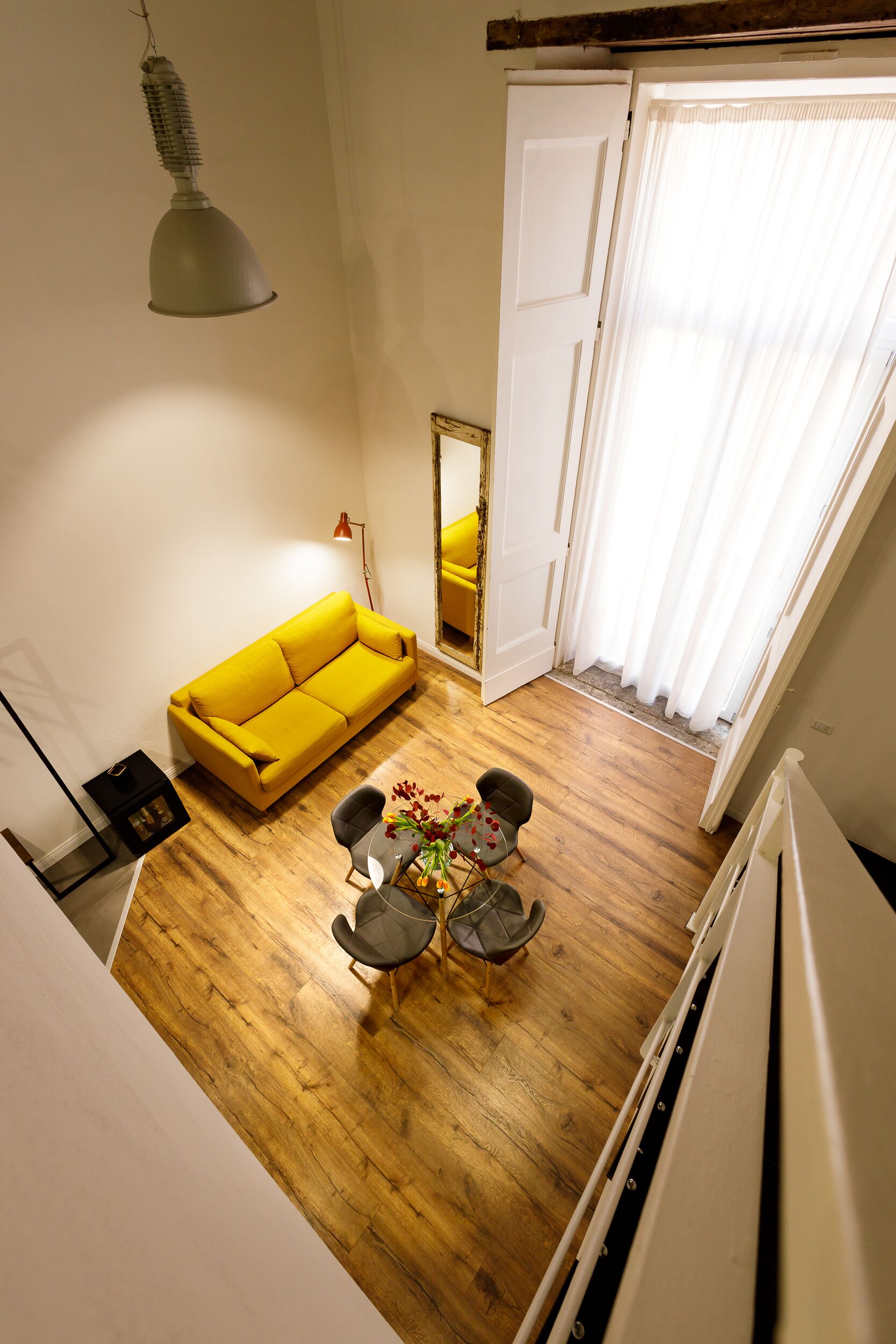 A living room with a yellow sofa, a small round table with chairs, a hanging lamp, and a large mirror on the wall.