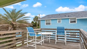 Een houten terras met twee stoelen en een tafel, een blauw huis op de achtergrond, en palmbomen.