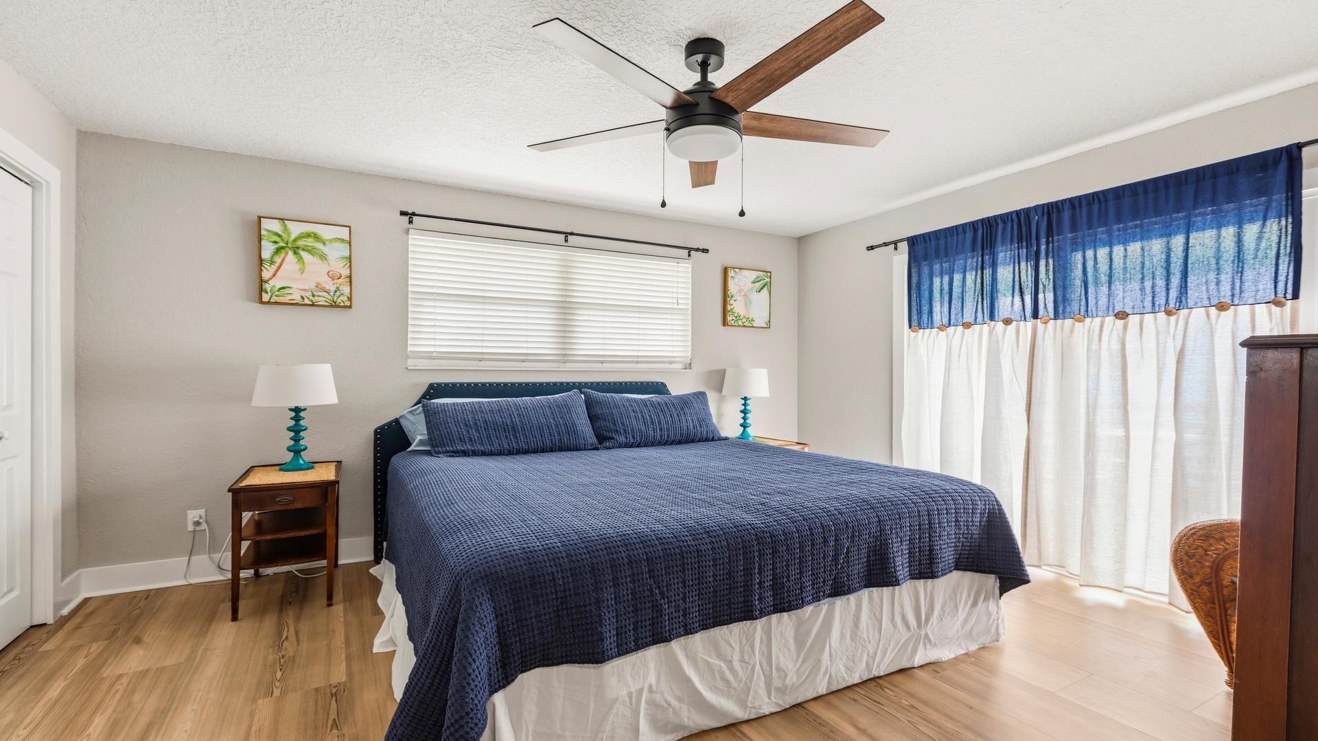 A bedroom with a bed, two bedside tables, a ceiling fan, a window with blinds, and curtains.