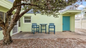 Un petit espace extĂ©rieur amĂ©nagĂ© avec deux chaises bleues et une table, un arbre, et un bĂątiment dotĂ© dâune porte et de fenĂȘtres vertes.