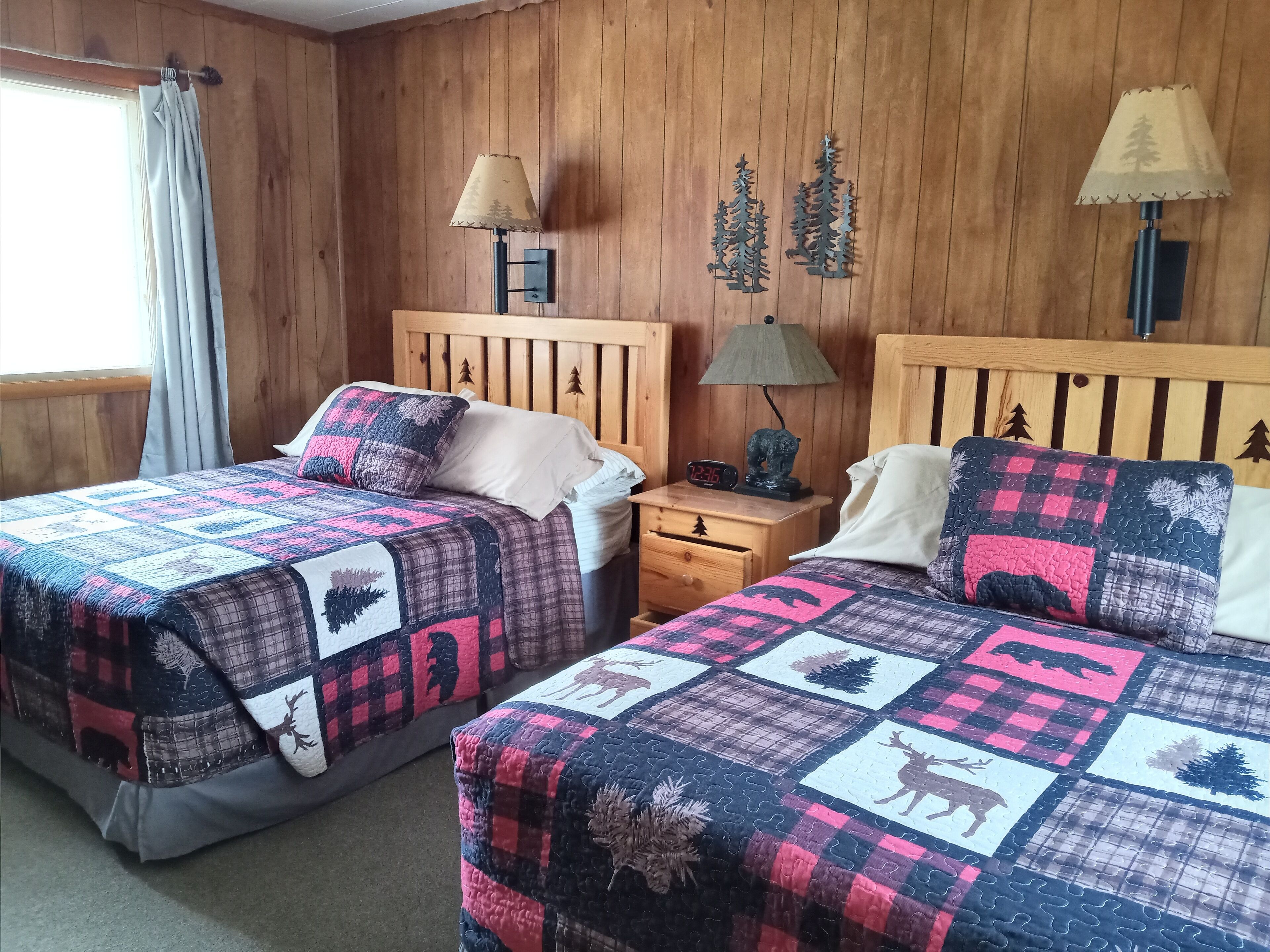 A room with two beds, each with a quilted bedspread featuring animal prints. There are two wall-mounted lamps with tree designs, a nightstand with a clock, and a window with curtains.