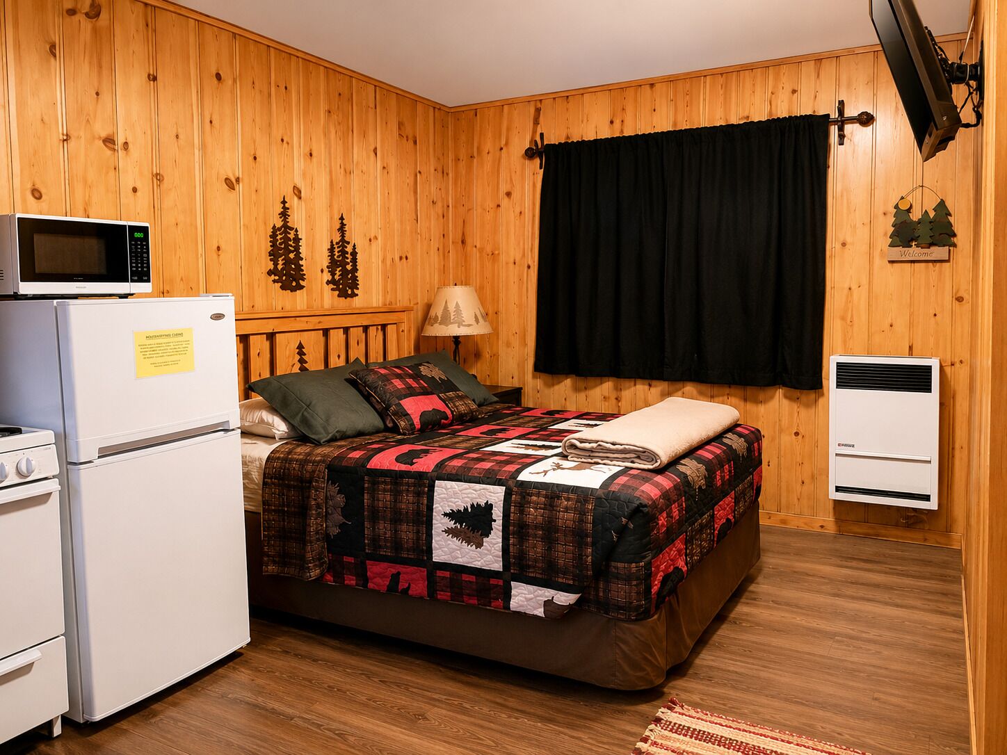 A wooden-paneled room with a bed, a microwave, a refrigerator, a heater, and a window with a black curtain.