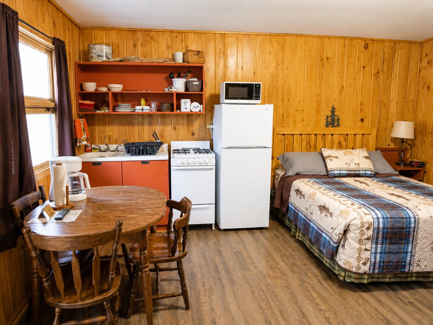 A compact room with a wooden interior, a bed with patterned bedding, a small kitchenette with a microwave, and a dining area with a table and chairs.