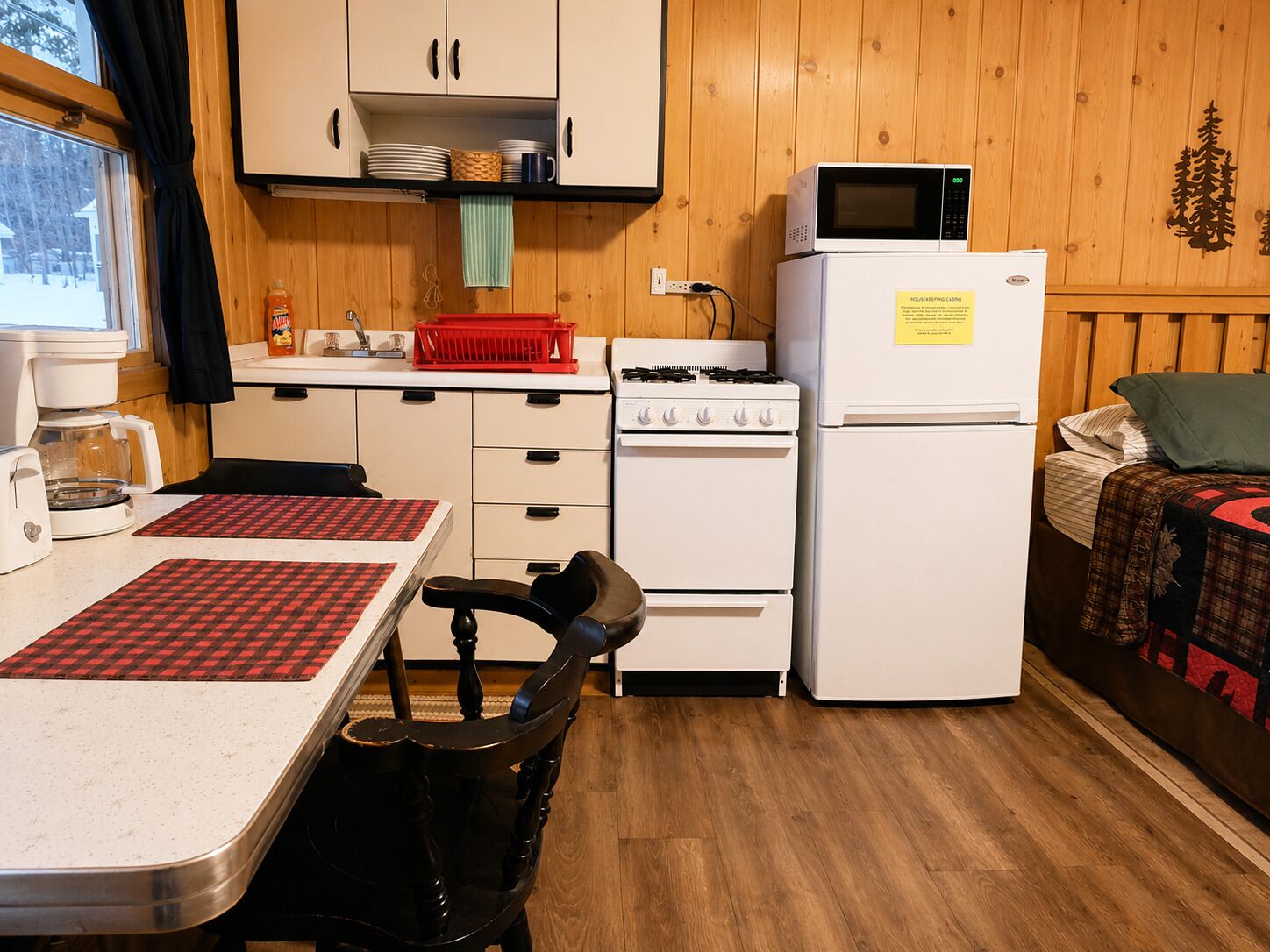 A compact kitchen with a stove, microwave, and refrigerator, a small dining table with a checkered placemat, and a bed with a quilt.
