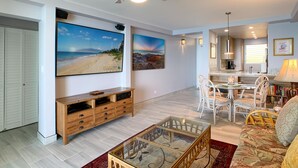 Un vaste espace de vie agrémenté d’un ventilateur de plafond, d’un grand téléviseur à écran plat, d’un meuble de rangement en bois, de meubles en osier et d’œuvres d’art représentant une plage et un paysage désertique.