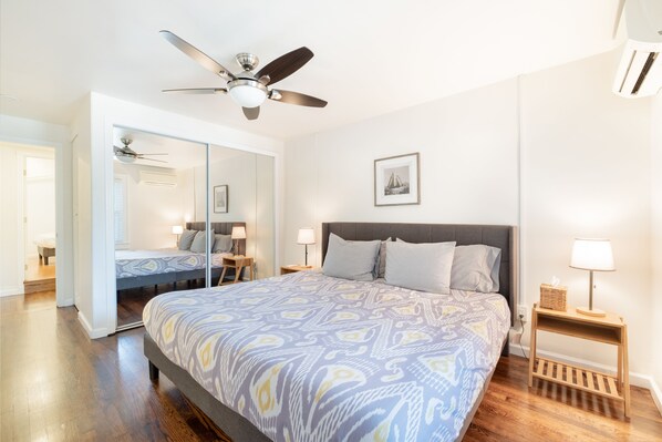 Un dormitorio con cama, ventilador de techo, mesita de noche con lámpara, estante en la pared y un cuadro enmarcado.