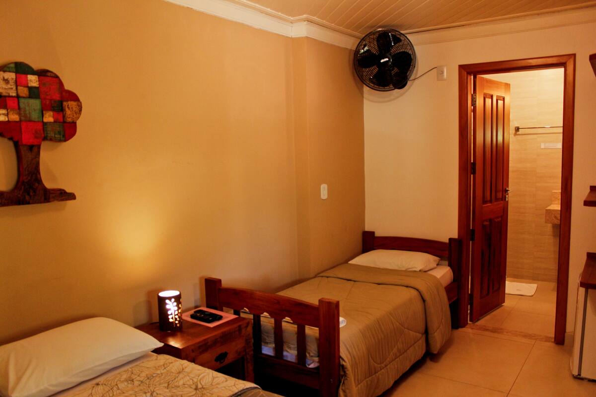 Un dormitorio pequeño y acogedor con dos camas individuales, una mesita de noche con lámpara, un ventilador en la pared y una puerta de madera que da a un baño.