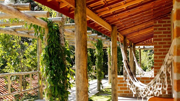 A covered outdoor area with wooden beams and brick walls, featuring a hammock and a bench. - Pousada Araucária (São Luiz do Paraitinga)