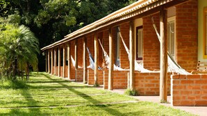 A row of brick buildings with hammocks hanging from the roof, surrounded by green grass and trees. - Pousada Araucária (São Luiz do Paraitinga)