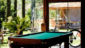 A pool table in a room with large windows, a bicycle, and a view of a garden and a building with a tiled roof. - Pousada Araucária (São Luiz do Paraitinga)