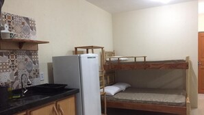 Cocina compacta con refrigerador, lavamanos y armarios de madera. Una cama alta está ubicada a la derecha de la cocina.