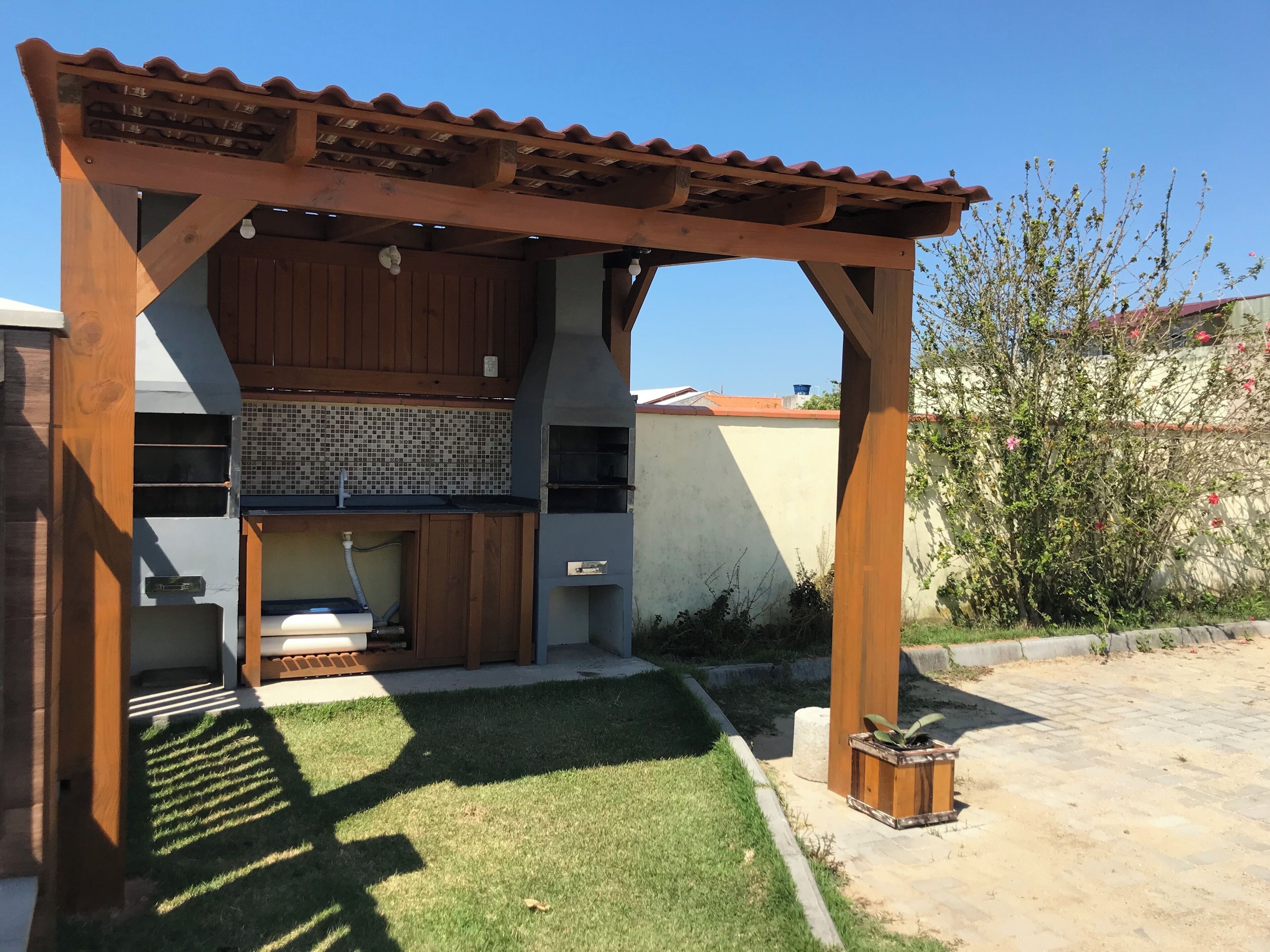 Una pergola de fusta amb teulada de rajoles, una barbacoa integrada i un petita rentaplat.