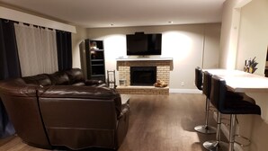 Un salon avec un canapé en cuir, une cheminée, un téléviseur et un coin bar équipé de tabourets.