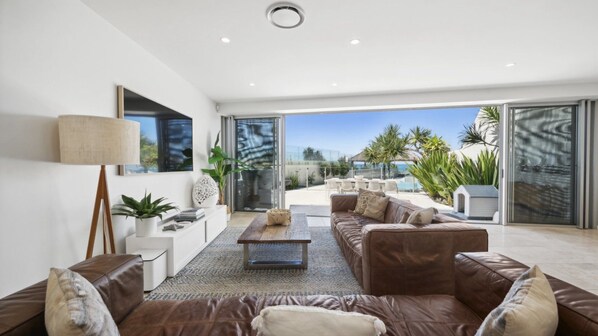 A modern living room with a brown leather sofa, a coffee table, and a large window offering a view of a pool and palm trees.