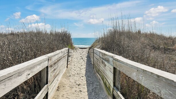 Un sentier en bois qui mène à la plage, avec le sable et la mer en arrière-plan.