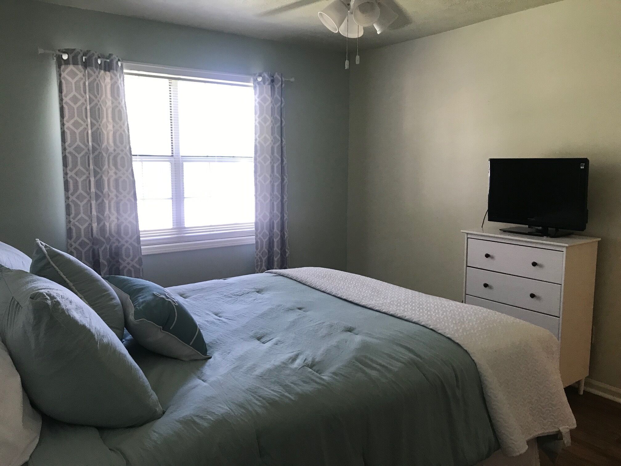 A bedroom with a bed, pillows, a dresser, a television, and a window with curtains.