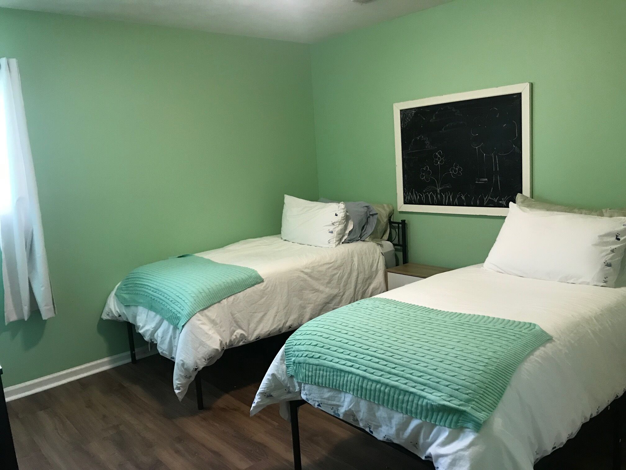 Una habitación con dos camas individuales, cada una con colcha verde y blanca, y una cortina blanca a la izquierda.
