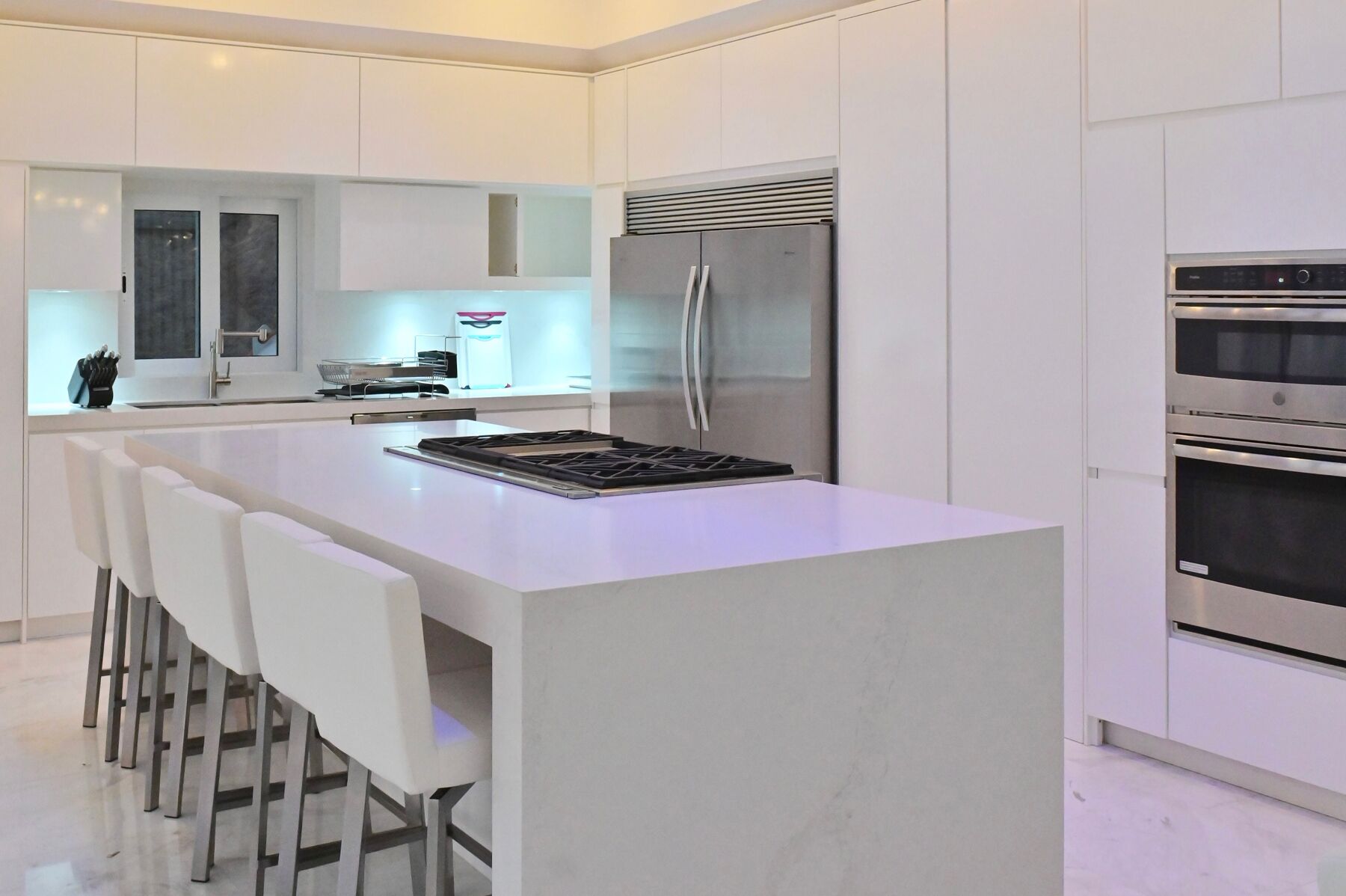 A modern kitchen with white cabinetry, a central island with bar stools, a stainless steel refrigerator, and built-in ovens.