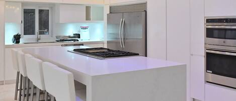 A modern kitchen with white cabinetry, a central island with bar stools, a stainless steel refrigerator, and built-in ovens.