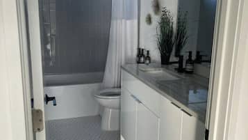 A modern bathroom with a hexagonal tile floor, a white toilet, a bathtub with a shower curtain, and a vanity with a mirror and toiletries.