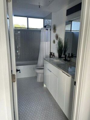 A modern bathroom with a hexagonal tile floor, a white toilet, a bathtub with a shower curtain, and a vanity with a mirror and toiletries. - Luxury Penthouse 2 Blocks | Near UT Stadium (Austin)