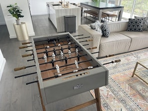 Uma mesa de futebol de mesa em uma sala de estar moderna, com um sofá e uma área de jantar ao fundo.