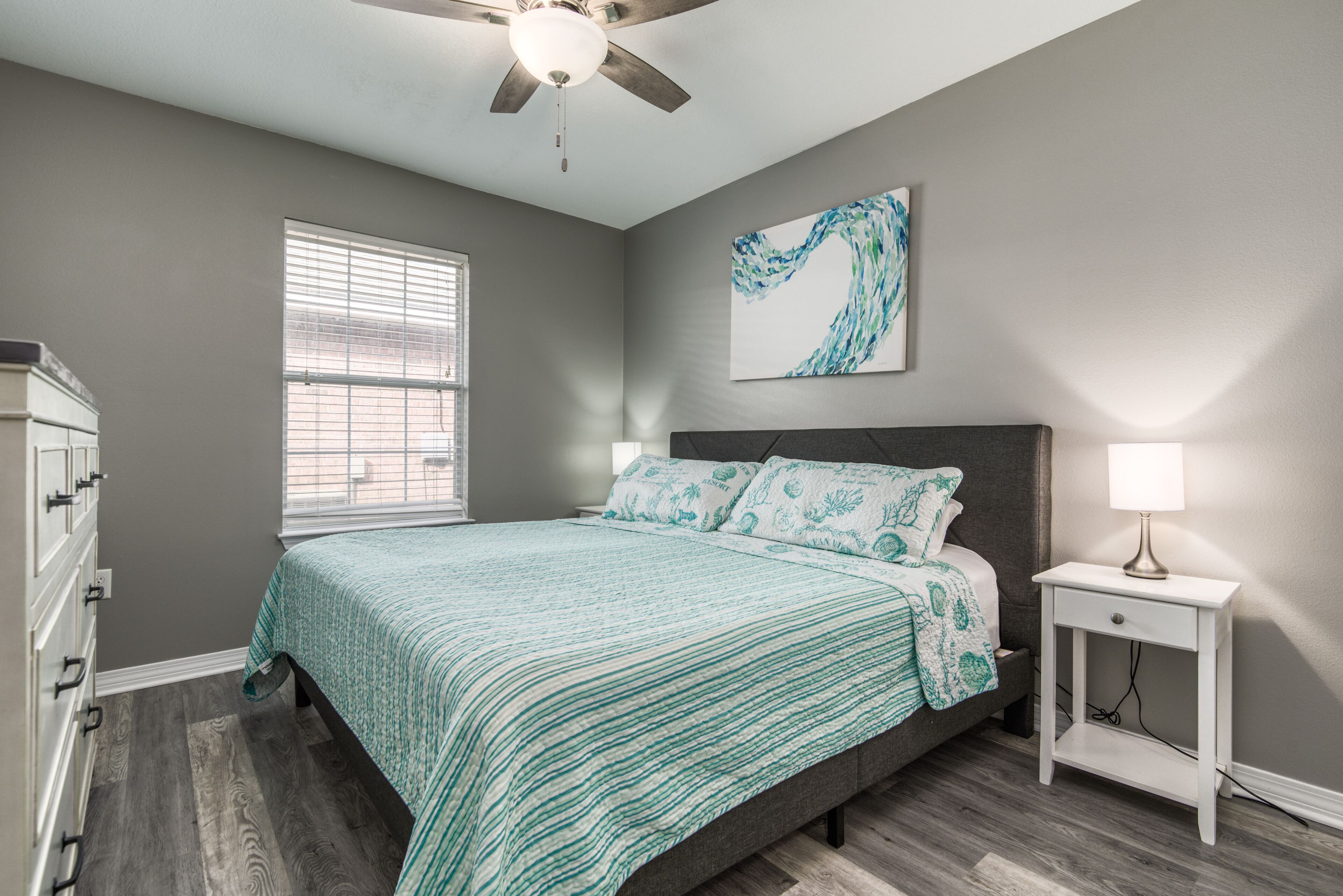 A bedroom with a bed, a nightstand, a ceiling fan, a window, and a wall-mounted artwork.