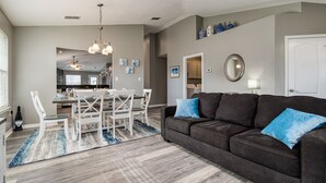 A modern living room with a dark brown sofa, blue cushions, and a dining area with white chairs and a table. - Private Pool! Walk to Beach! 3 Kings & Quad Bunk! (Miramar Beach)