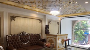 Une chambre luxueuse avec un canapé marron à motifs capitonnés, une table basse en verre, des décorations dorées élaborées au plafond et une vue sur l’extérieur grâce à une grande fenêtre.