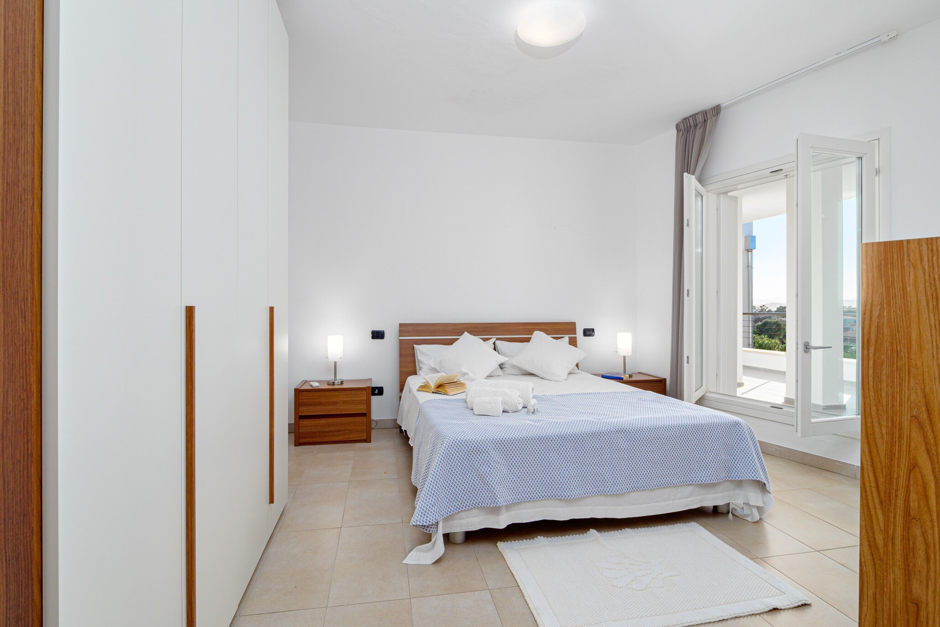 A bedroom with a bed, bedside tables, a nightstand, a window with a view, and a door leading to a balcony.