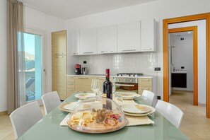 A modern dining area with a glass table, white chairs, a wine bottle, and a view through the glass door.