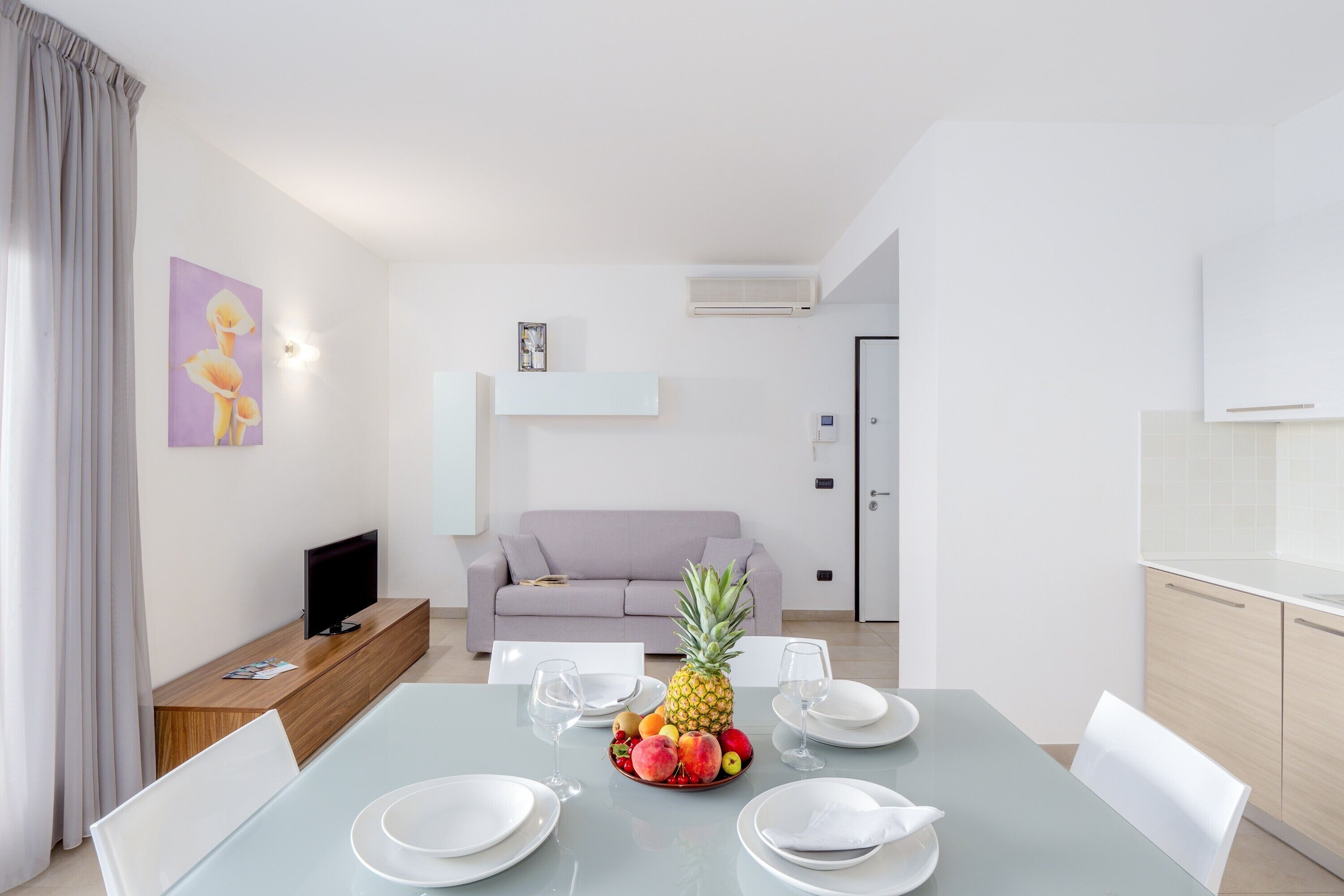 A modern dining area with a table set for four, a fruit bowl, a sofa, and a TV in the background.