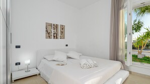 A modern bedroom with a large bed, a bedside table with a lamp, and a window with a view of greenery. - Spacious Apartment "Villa Marconi 6" with Sea View, Private Terrace & Garden (Golfo Aranci)