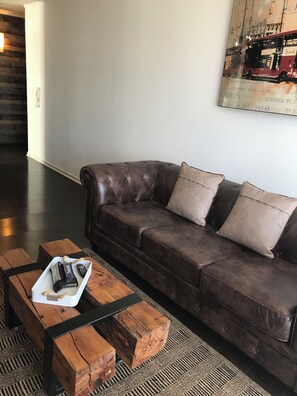 Un sofá de cuero marrón oscuro con tres cojines, una bandeja blanca con un control remoto y una mesa de centro de madera con una alfombra.