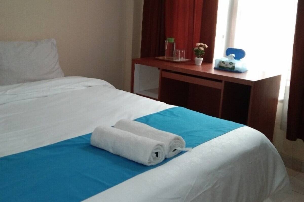 Een net opgemaakt bed met wit en blauw beddengoed, een houten nachtkastje met een vaas en een kleine plant, en een raam met rode gordijnen.